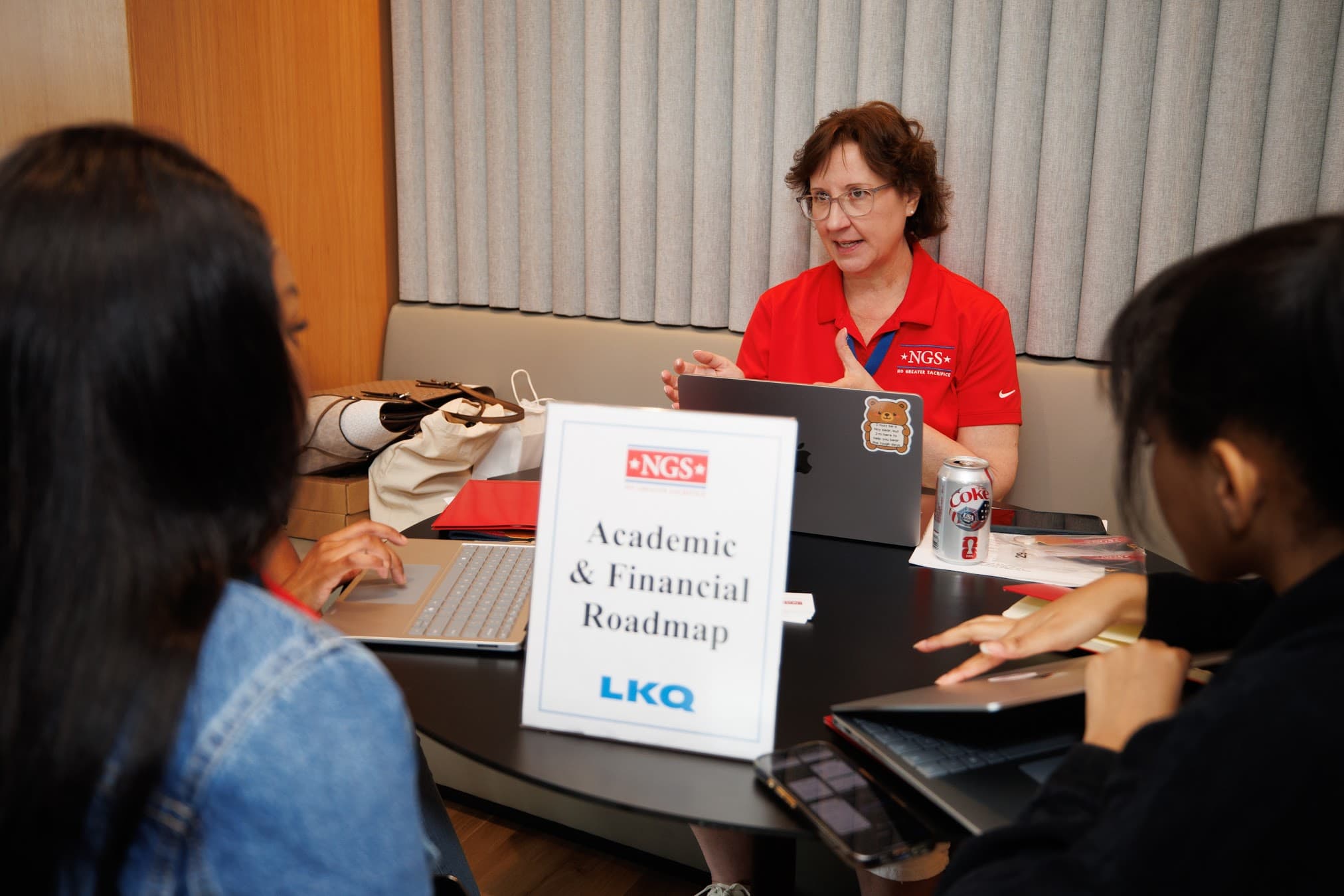 An NGS staff member in a red NGS polo talks with Scholars around a table during a 2026 BootCamp session, beside a sign reading “Academic & Financial Roadmap”.