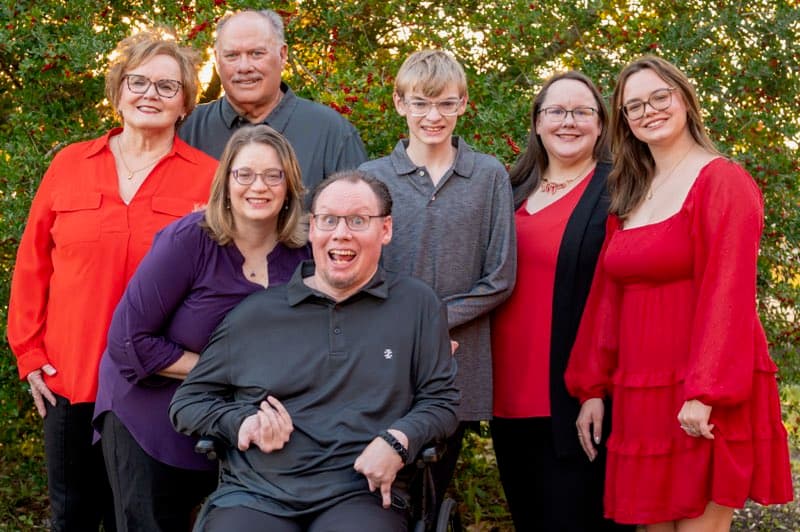 The Edmundson family poses in red and dark tones before a holly hedge: Eric seated in a wheelchair at center, surrounded by Stephanie, Hunter, Gracie, Anna, Beth, and Edgar.
