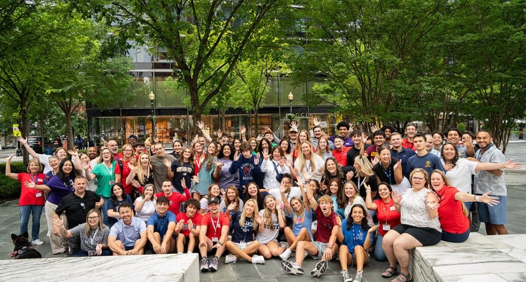 Three rows of NGS Scholars in college shirts - including UCF, Arizona State, South Alabama, Ole Miss, and Rogers State - pose beneath a red, white, and blue balloon arch in front of an NGS backdrop.