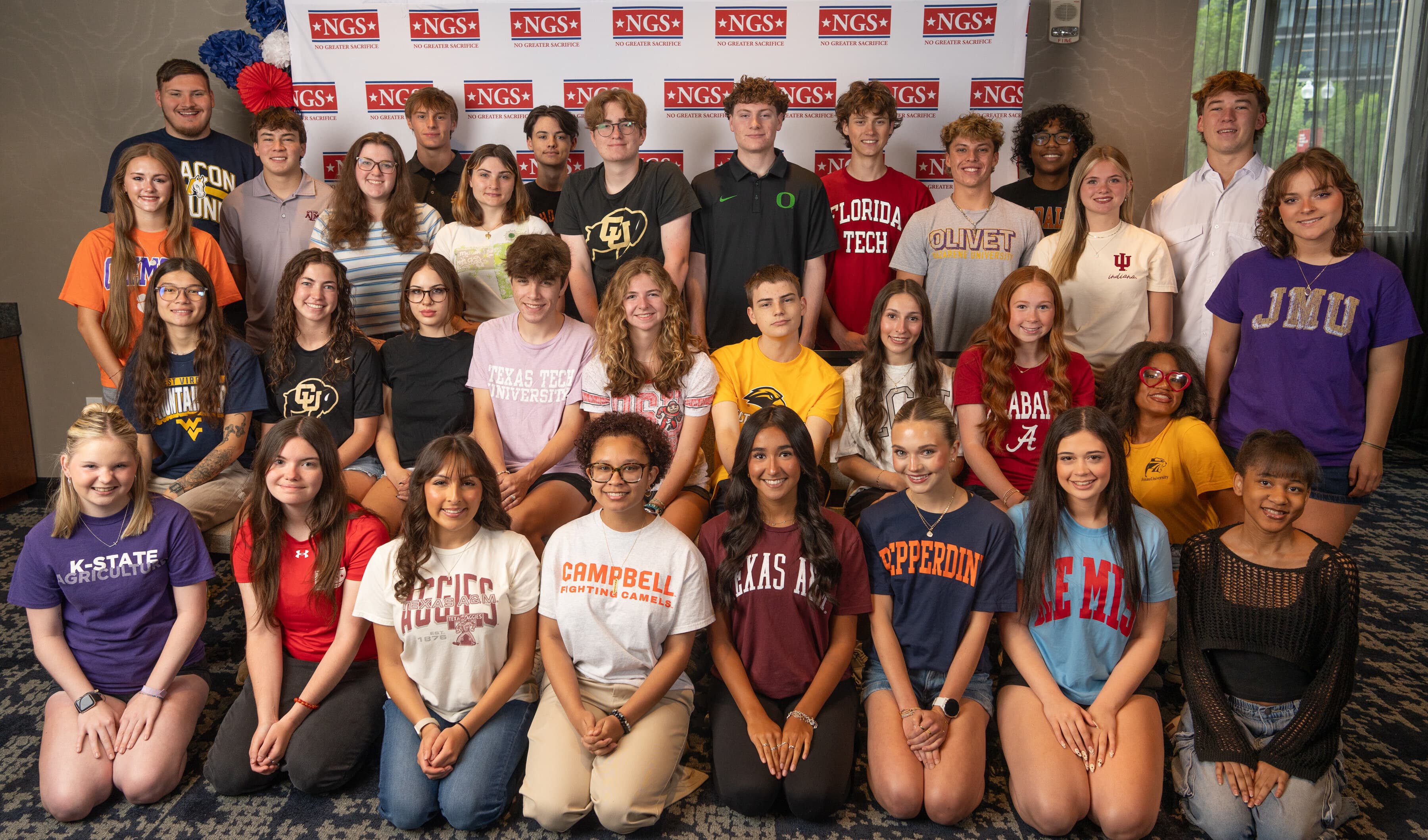 Three rows of NGS Scholars in college shirts, including Texas Tech, Florida Tech, JMU, K-State, Campbell, Texas A&M, Pepperdine, and Ole Miss, pose in front of an NGS backdrop.