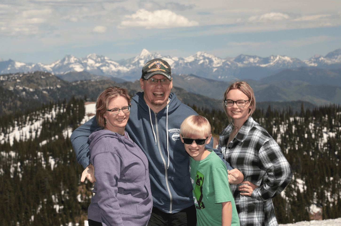 Stephanie, Eric, Hunter, and Gracie Edmundson hug and grin on a snowy mountain overlook with jagged peaks behind them.