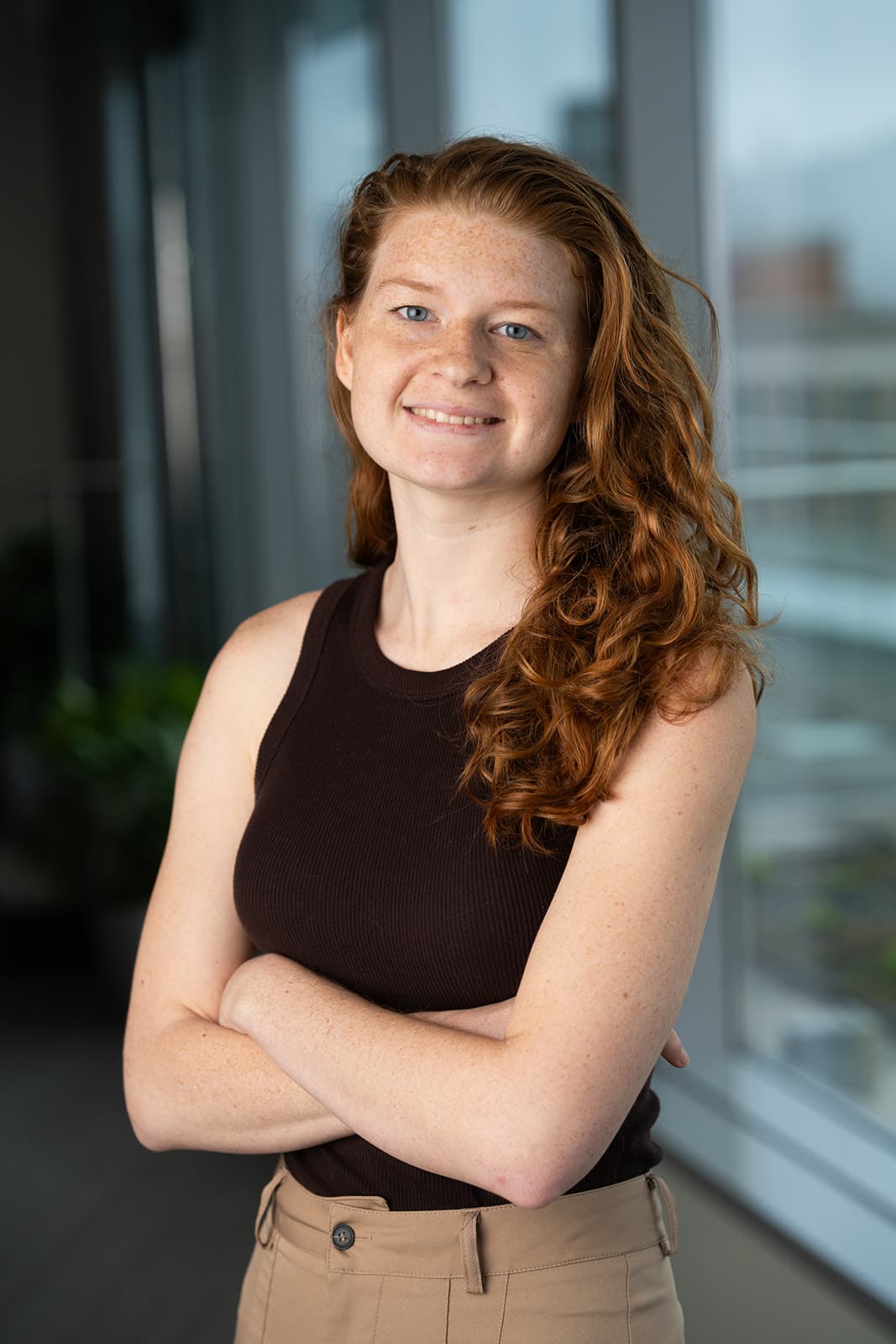 Paiten Prescott, a young woman with long curly red hair, smiles with arms crossed in a dark sleeveless top beside a window overlooking the city.