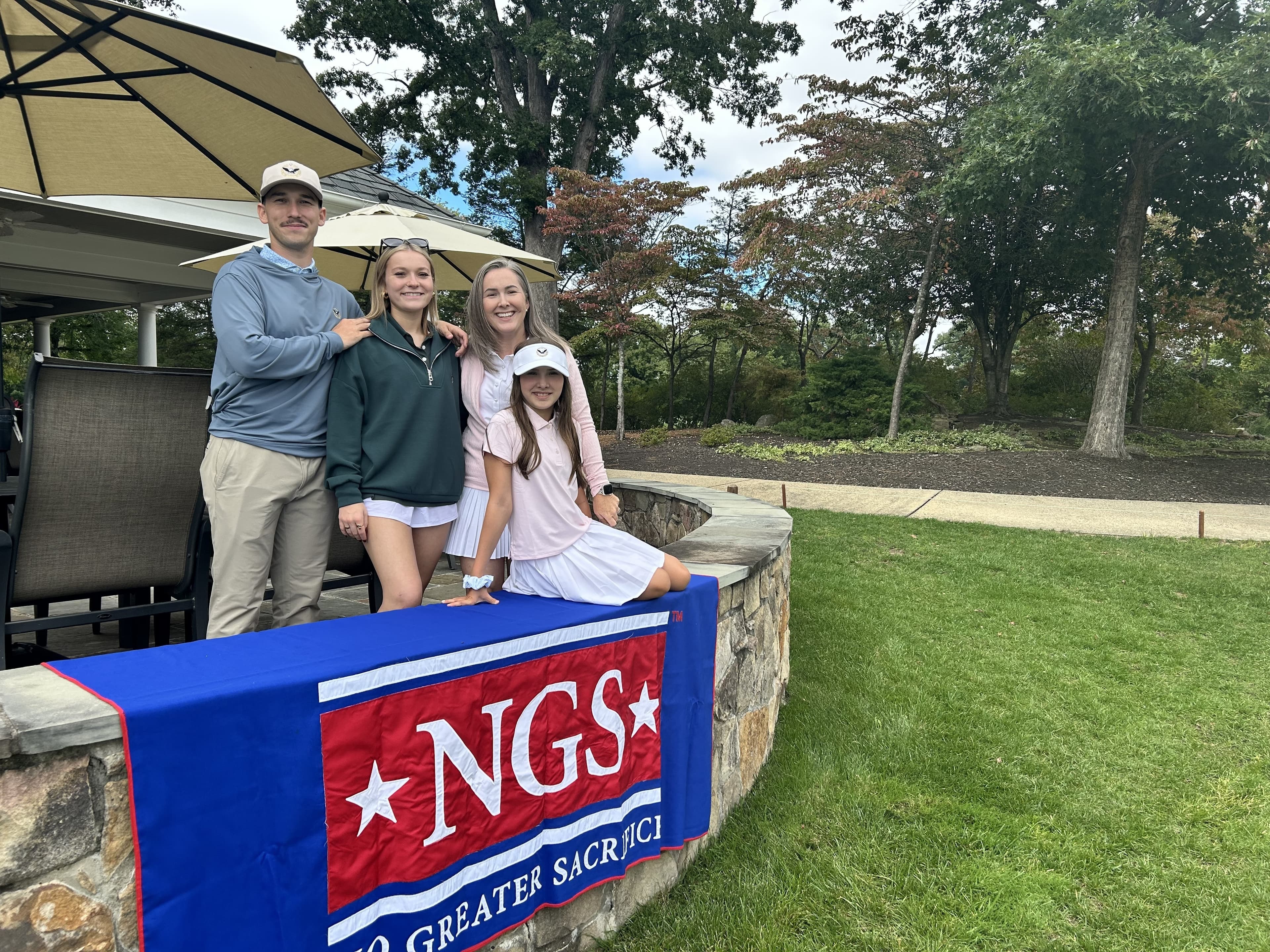 Four members of the Chism family - a young man in a cap, two young women, and their mother - smile behind a stone wall draped with an NGS banner on the golf course patio.