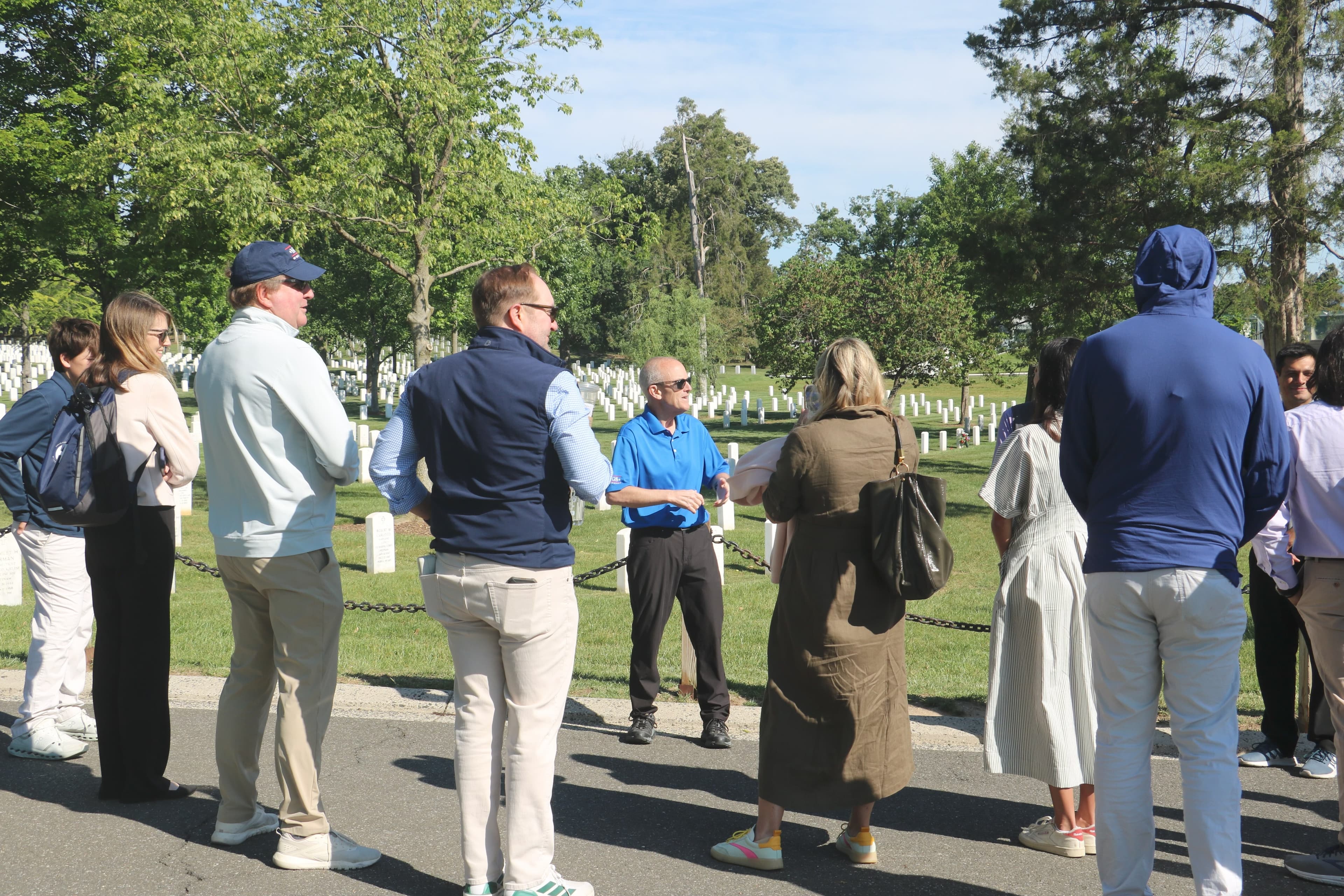 A group of NGS supporters stands along a chain fence listening to a man in a blue polo speak, rows of white headstones and green trees behind him at Arlington National Cemetery.