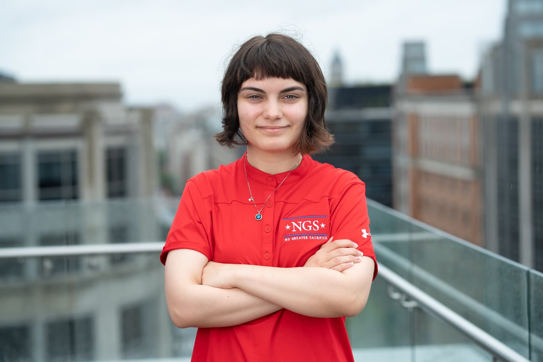 Kristian Engeldrum, a young woman with short dark hair, smiles with arms crossed in a red NGS polo on a rooftop terrace with city buildings behind her.