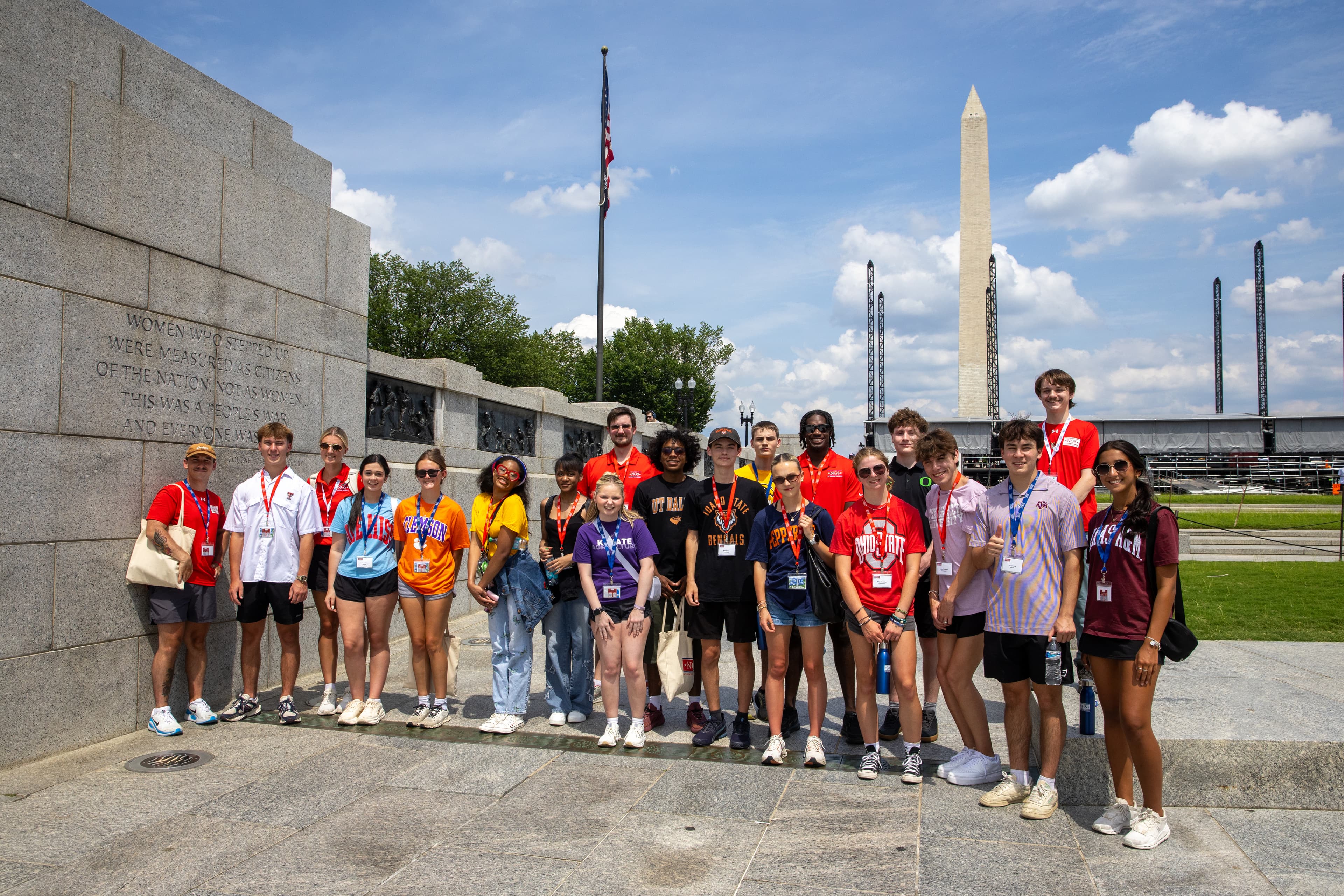 About twenty NGS Scholars in college T-shirts pose with NGS staff at the World War II Memorial on a sunny day, the Washington Monument rising behind them.
