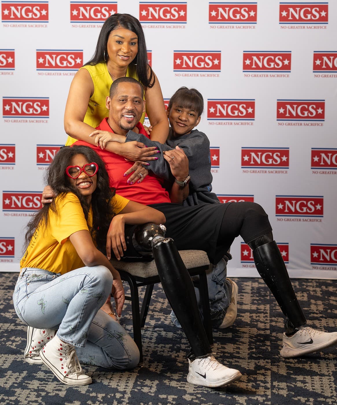 The King family in front of an NGS backdrop: Khieda King standing behind her husband Cedric, a double amputee seated in a red shirt with prosthetic legs, flanked by daughters Amari, kneeling in heart-shaped sunglasses, and Khamya, leaning in with an arm around him.