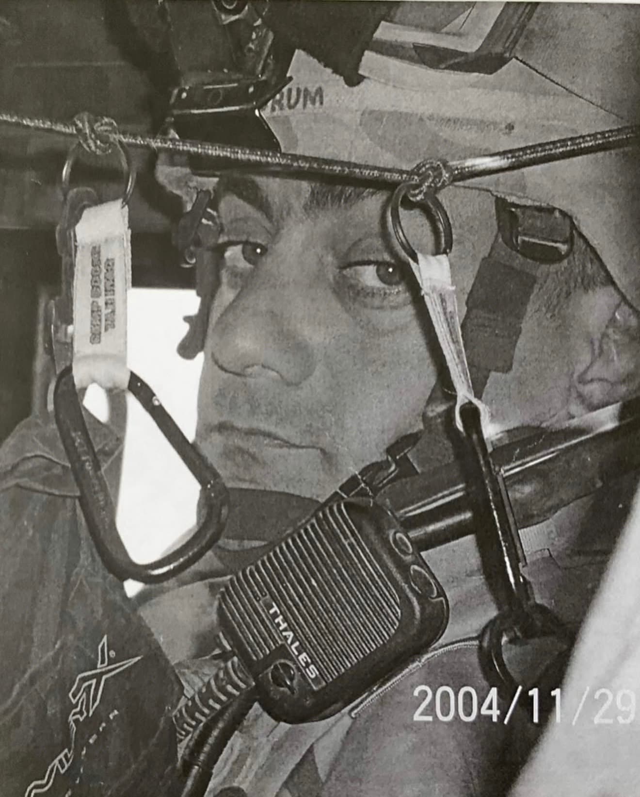 Black-and-white photograph dated 2004/11/29 of Army SSG Christian Engeldrum in helmet and body armor inside a military vehicle, a radio handset and carabiners hanging in the foreground.
