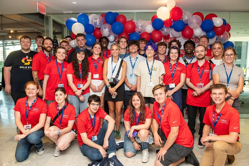 About thirty NGS Scholars, most in red NGS polos with lanyards, pose beneath a red, white, and blue balloon arch.