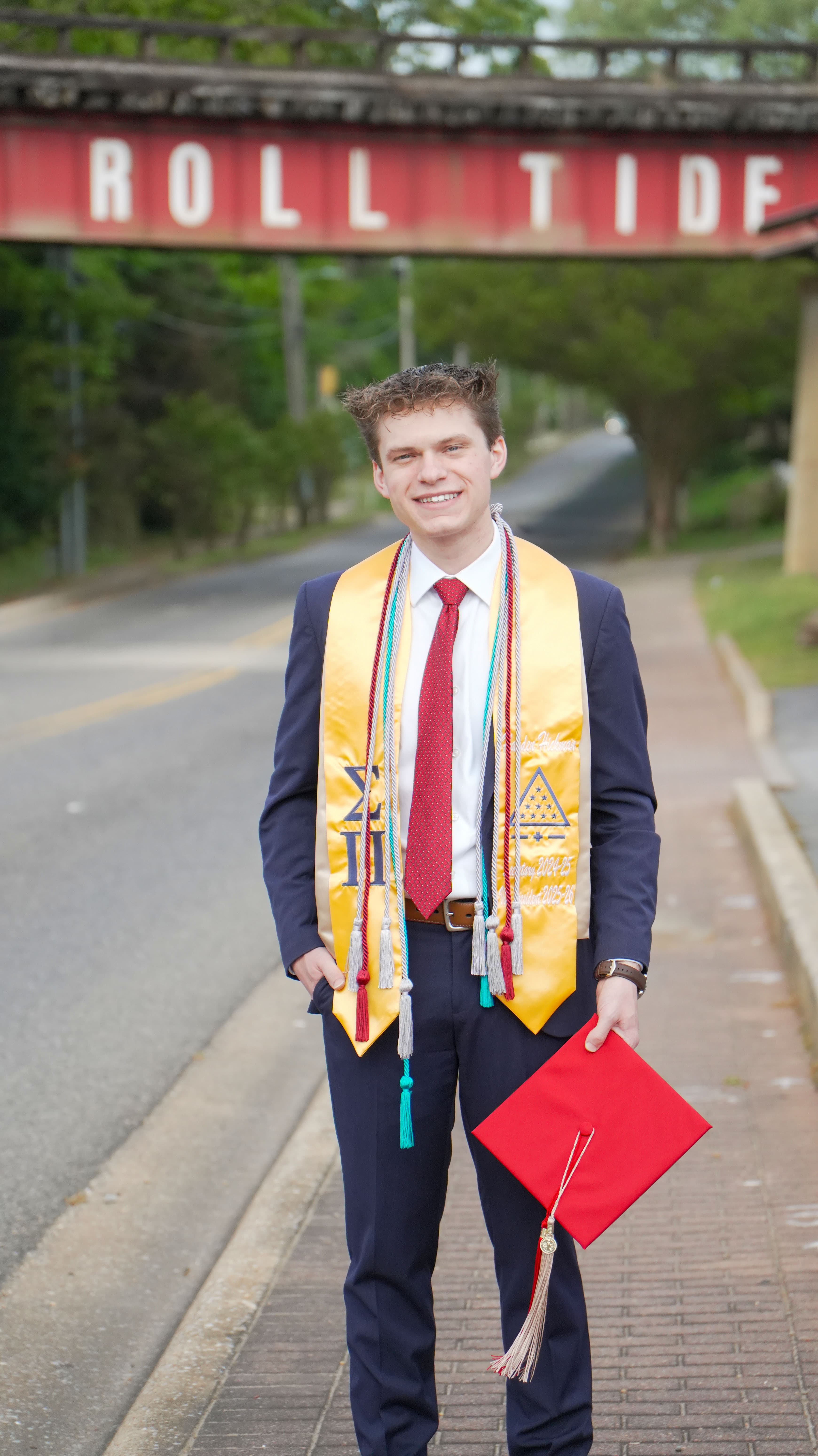 Hayden Hickman in a navy suit, red tie, gold honor stole, and cords holds a red mortarboard beneath a railroad bridge painted Roll Tide.