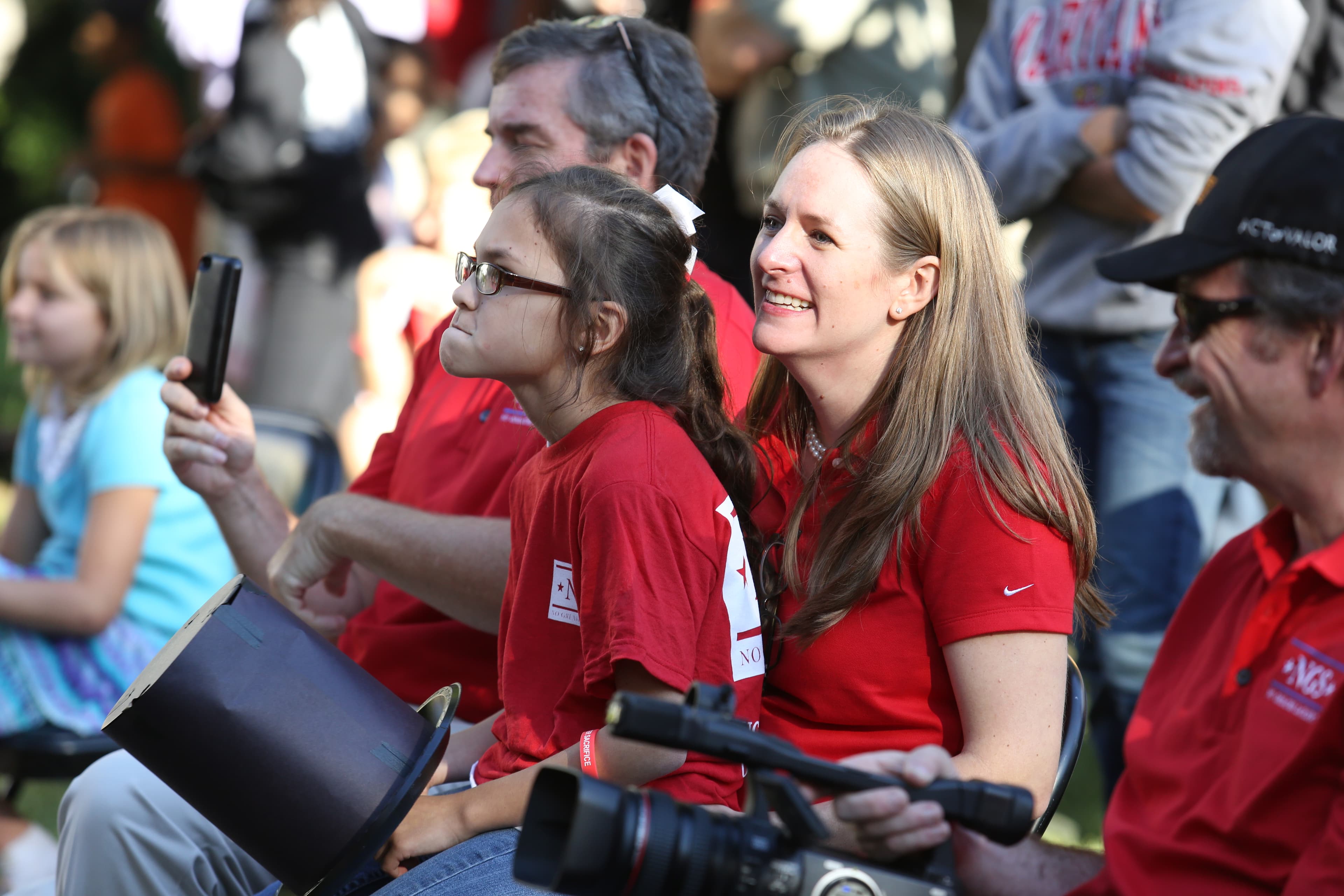 A smiling woman in a red Nike shirt sits alongside a young girl wearing glasses at the 2014 Freedom Award Family Day outdoor event. Both wear red NGS shirts and are engaged watching the program alongside other attendees.