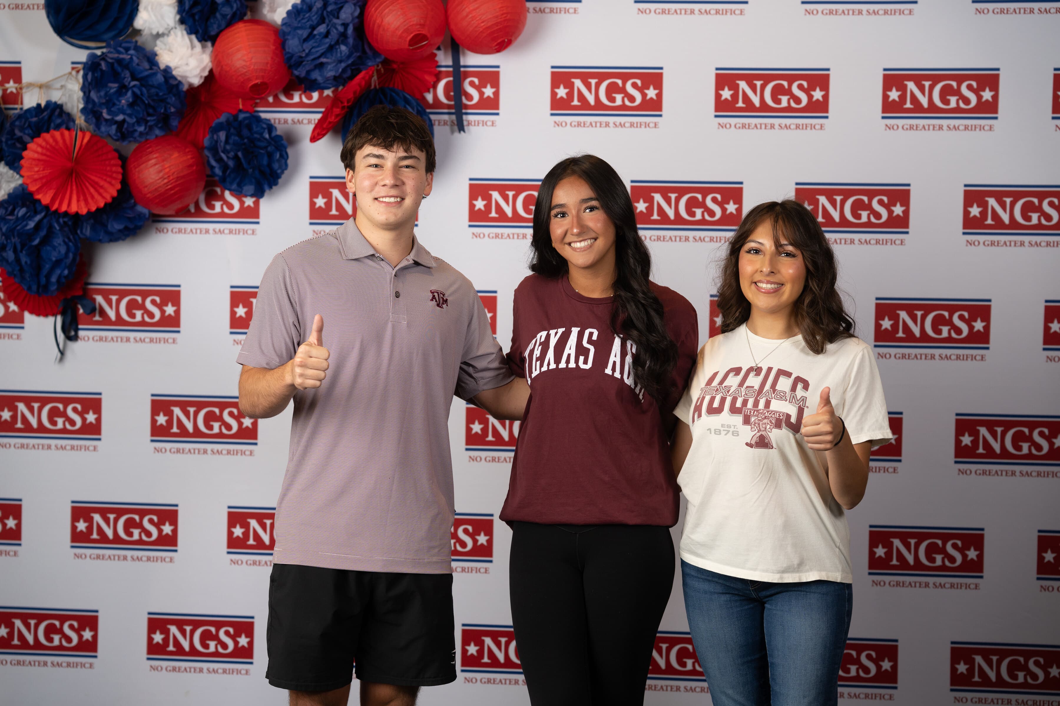 Three smiling Scholars in Texas A&M shirts give thumbs-up in front of an NGS step-and-repeat backdrop decorated with red, white, and blue paper lanterns.