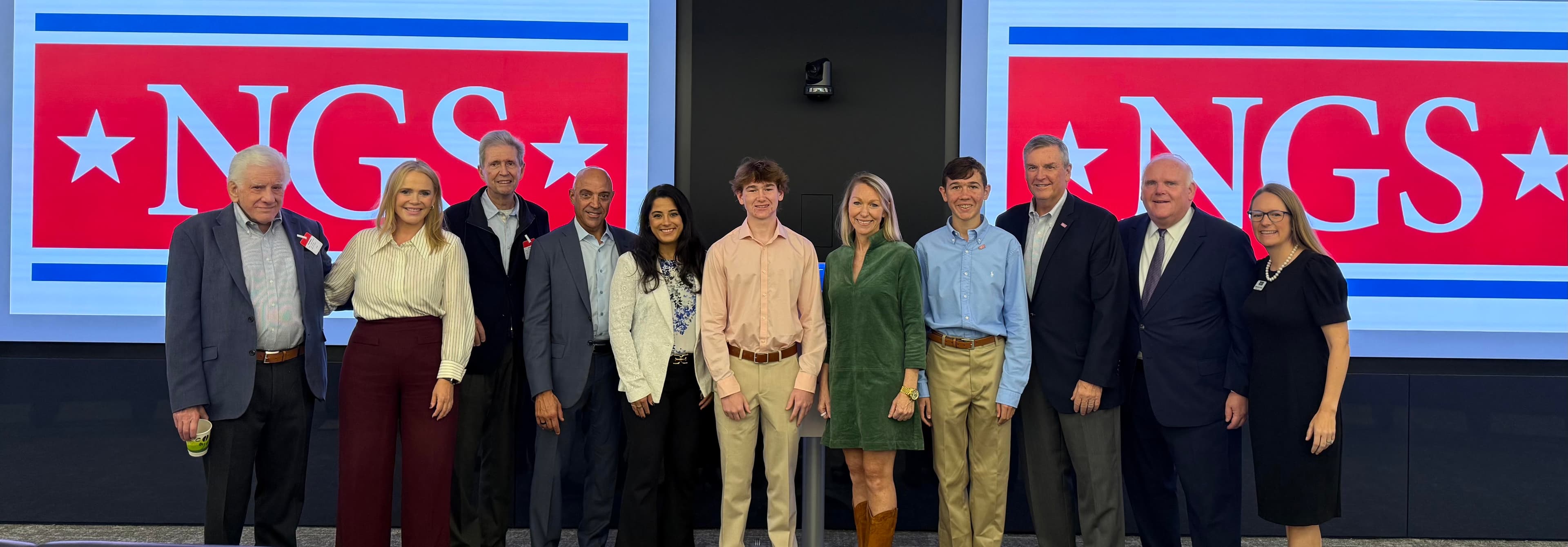 Eleven people, including two teenage boys and their mother, stand in a line on a stage between two large NGS logo screens at the UBS Manhattan Market kickoff.