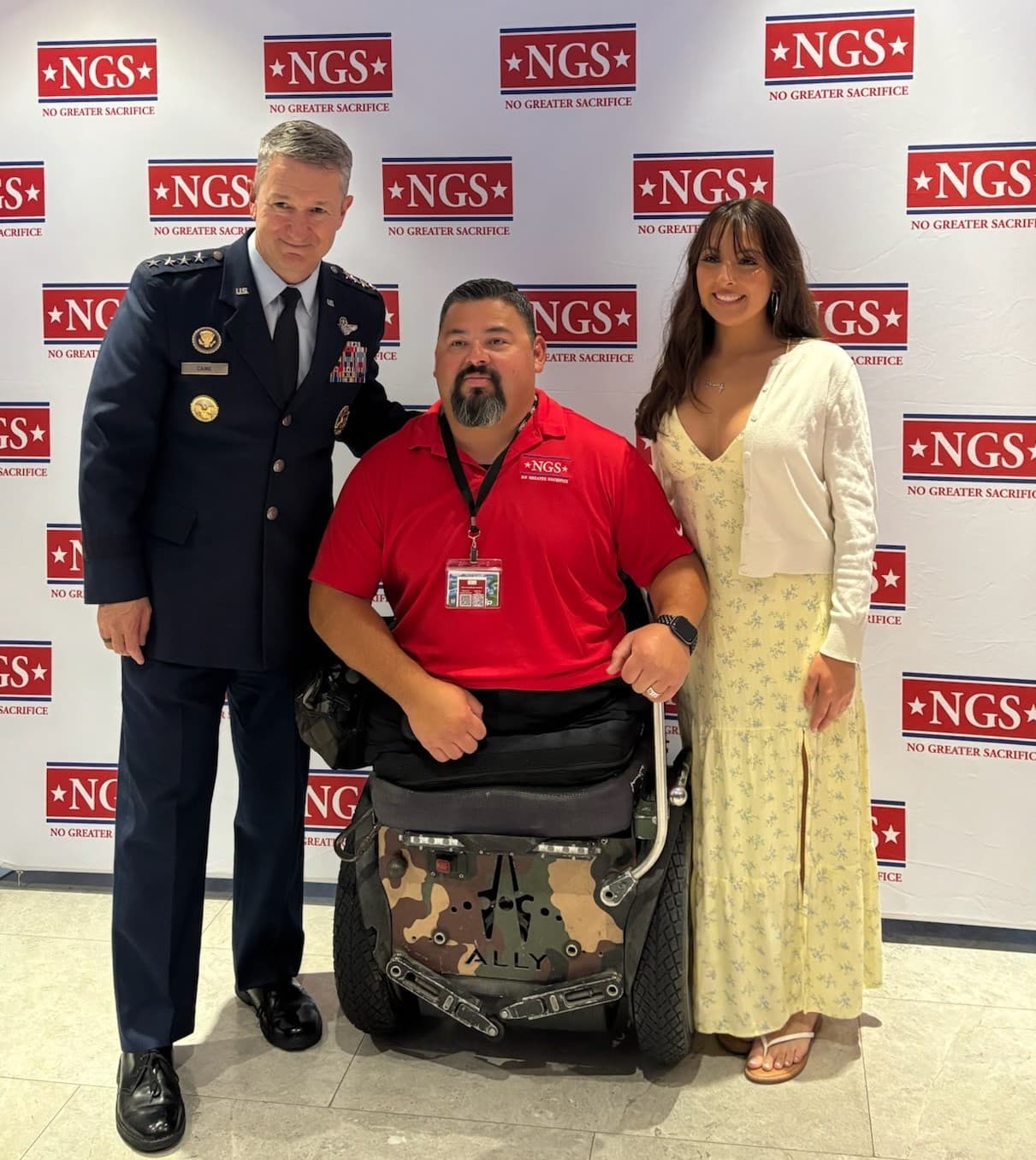 General Dan Caine in Air Force dress uniform with Gunnery Sergeant Guillermo Tejada, seated in a wheelchair, and NGS Scholar Aracely Tejada in front of a No Greater Sacrifice backdrop at the Freedom Award Reception.